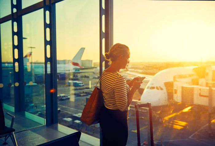 A young woman, absorbed in her cellphone, scowls as she gazes through the window of an airport departure lounge.