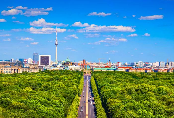 A highway flanked by lush forests leads to the vibrant metropolitan skyline, featuring a prominent TV tower in the distance.