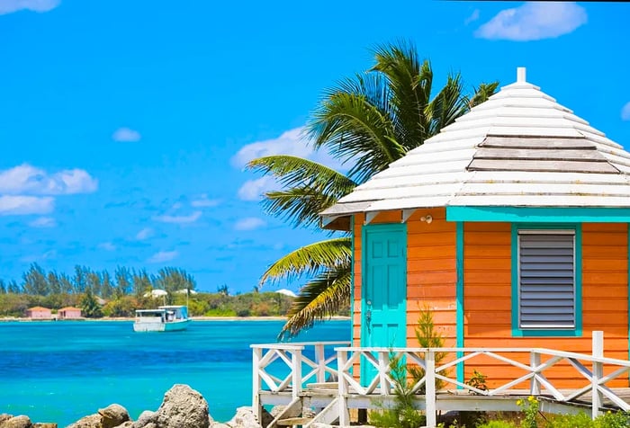 A beach hut perched on the rugged coastline overlooks turquoise waters and a solitary white sailboat.