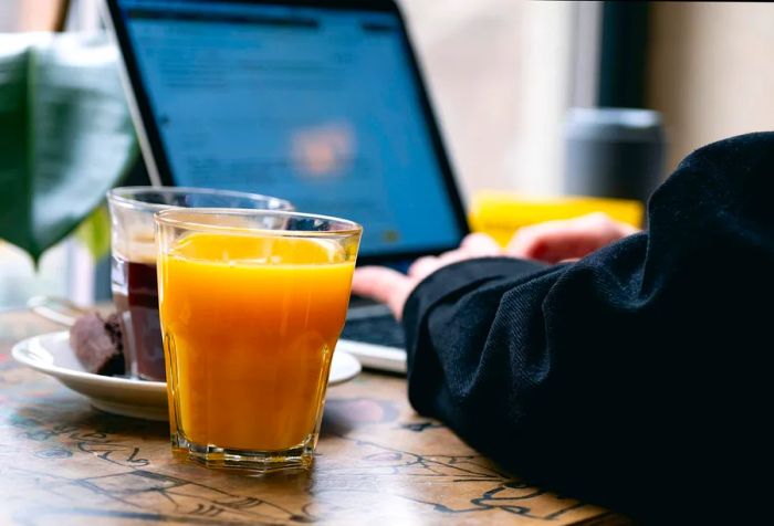 With great focus on their laptop, an individual works diligently, accompanied by a steaming mug of black coffee and a refreshing glass of orange juice placed on the table nearby.