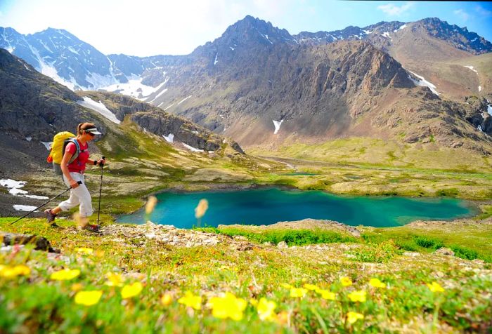 A female hiker strolls along the shore of a blue lake, framed by towering mountains.