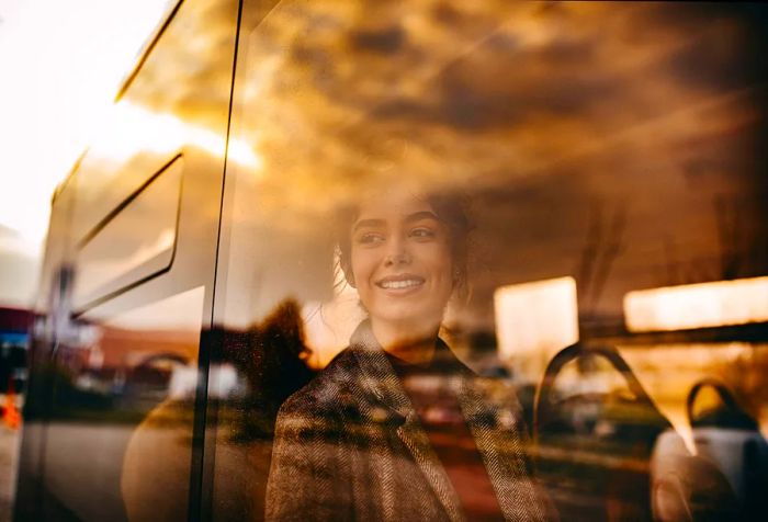 A woman in a coat beams as she gazes out the window of a bus.