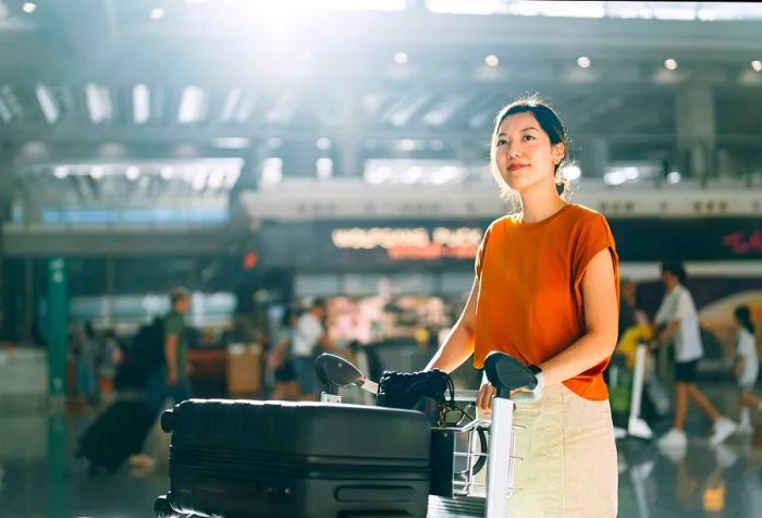 A young Asian woman is pushing a luggage trolley loaded with suitcases toward the check-in counter at the airport terminal, ready for her trip—a perfect blend of business travel and vacation.