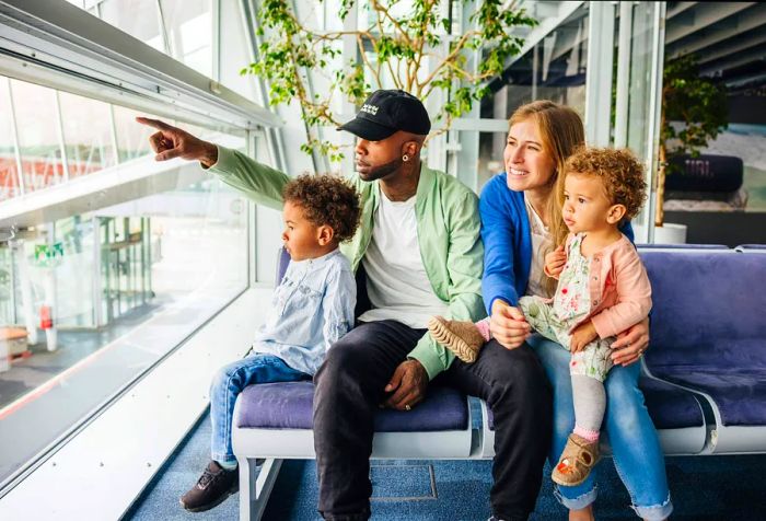 A family shares quality time in an airport lounge, with a parent pointing out something fascinating to the kids as they wait for their flight.