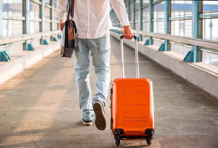 A casually dressed man, carrying a backpack and dragging a suitcase, makes his way along a pathway leading to a railway station.