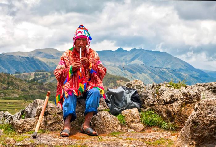 An elderly man adorned in a vibrant patterned poncho plays a musical instrument while seated on the rocks.