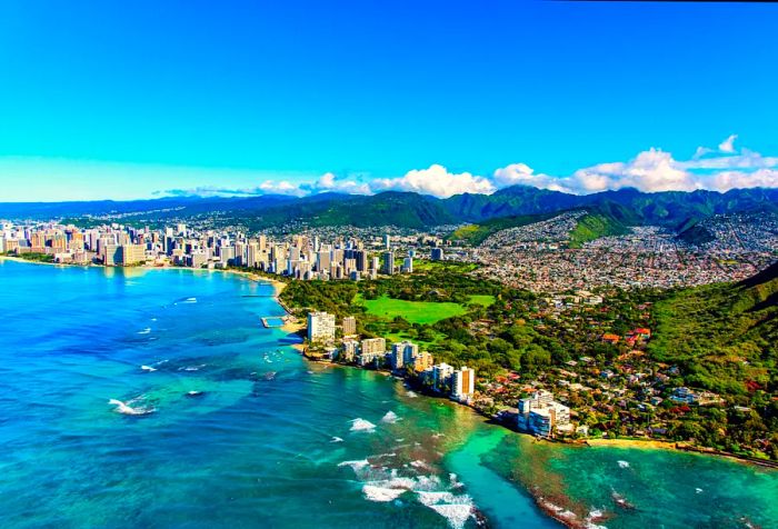 The entire coastline of Honolulu, Hawaii stretches from the base of Diamond Head crater and state park, flowing past the hotel-lined Waikiki Beach towards the distant downtown, with suburban neighborhoods nestled in the hills surrounding the city center.