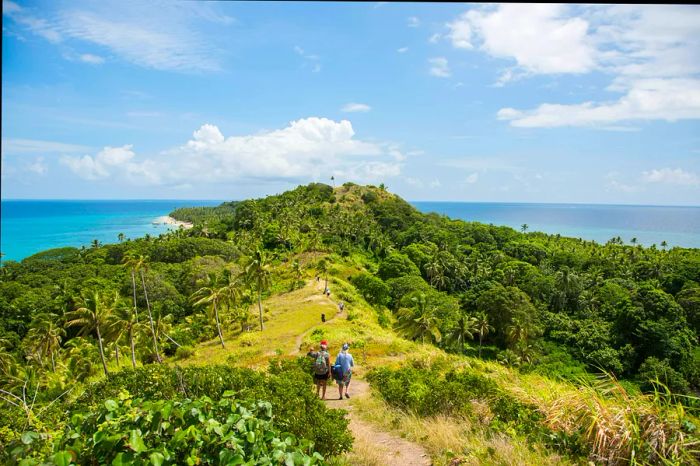 Tourists trek along a mountain ridge surrounded by lush greenery and the Pacific Ocean vista on Dravuni Island, Fiji