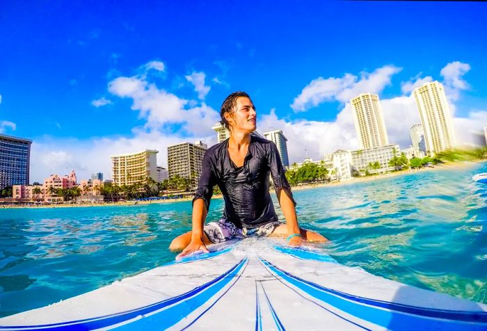 A person peacefully sits on a surfboard in the water, gazing into the distance, with buildings providing an urban contrast to the tranquil coastal scene.