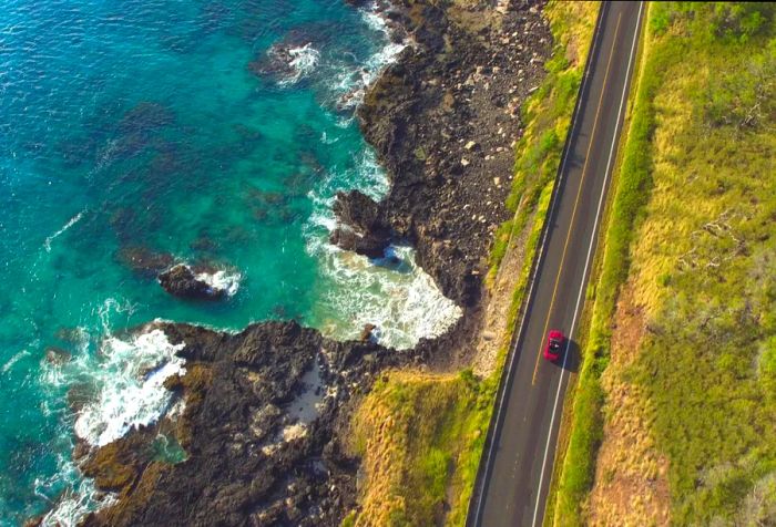 A vehicle drives along a coastal route beside a rugged beach where waves crash against the shore.