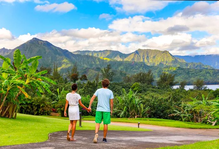 A lovely couple strolls hand in hand across a vibrant landscape, with a forested mountain range rising against a backdrop of a cloudy blue sky.