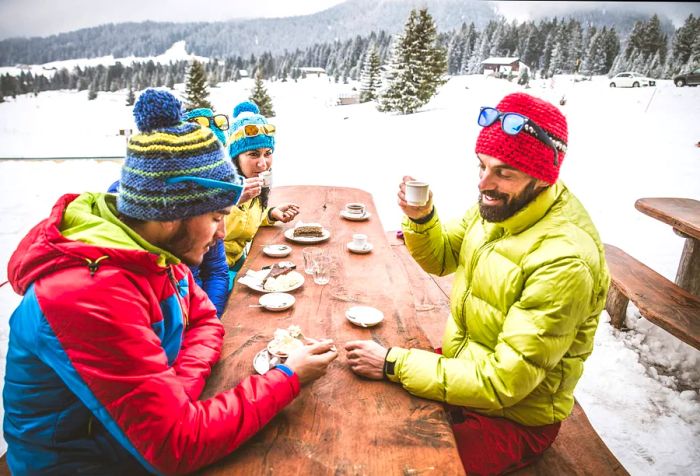 Three individuals bundled up for winter are seated at a wooden picnic table, relishing delicious food.