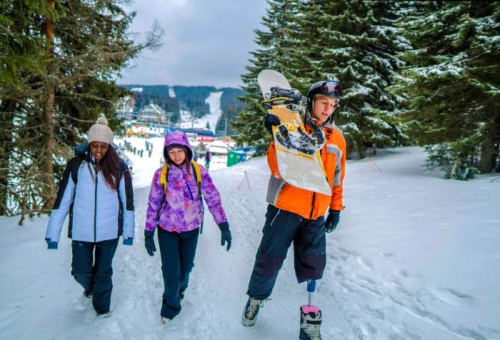 A man with prosthetic legs walks alongside two friends in a snowy landscape surrounded by trees.