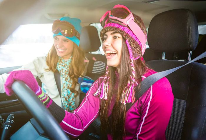 Two young women, clad in ski gear, are laughing in the front seats of a car.