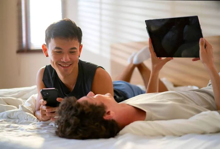 A couple relaxes on the bed, one reclining while the other leans comfortably against them, both smiling and sharing a digital device.