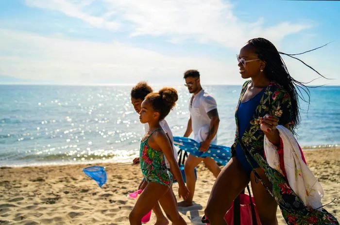 Parents and their children stroll along the beach, carrying a parasol and other beach essentials.