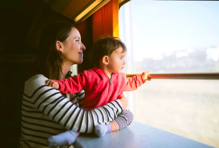 A mother wearing a striped sweater holds her baby as they gaze out the window of the train.