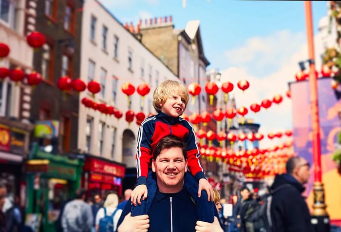 A man carries his son on his shoulders while walking down a street adorned with red lanterns overhead.