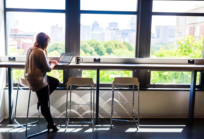 An employee is seated comfortably at a high table in a bright and spacious office pantry, working on her laptop.