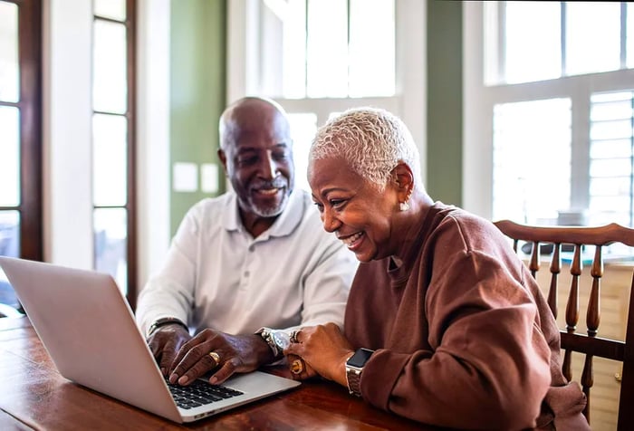 An elderly couple joyfully using a laptop on a wooden table.