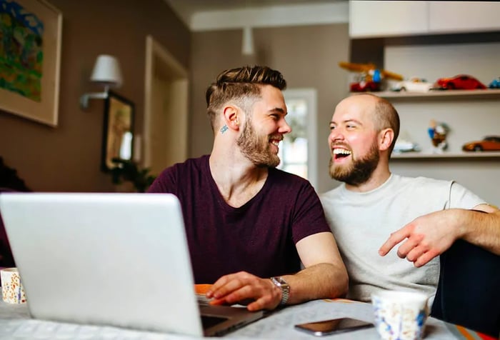 A cheerful gay couple sits in the dining area, sharing a laptop in front of them.