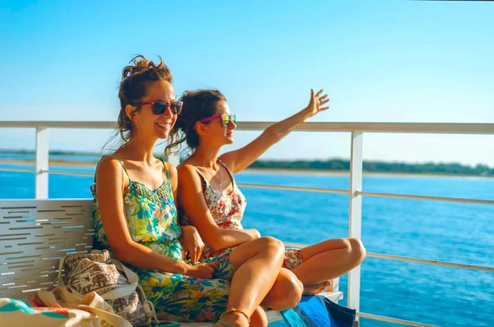 Two young women, either friends or sisters, sit on a bench on the ferry deck, waving to the horizon on a sunny summer day as they head to a tourist island destination.