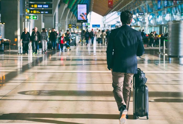 A rear view of a traveler with a suitcase and numerous other passengers walking through the airport corridor.