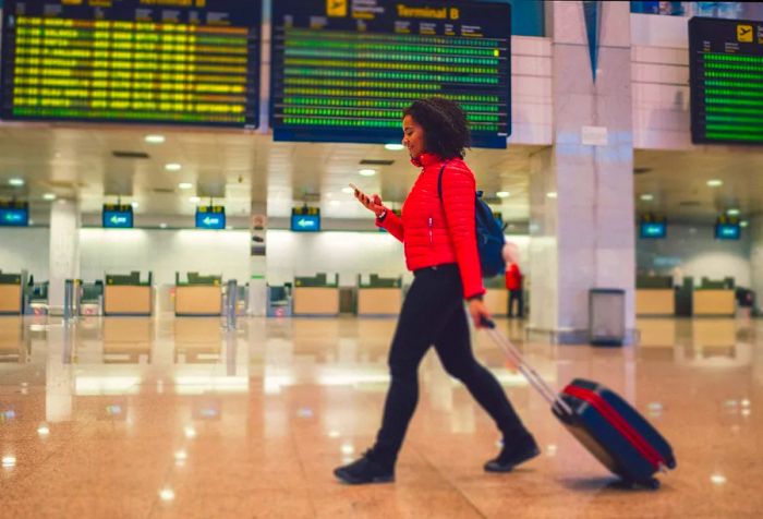 A woman dressed in a red jacket checks her phone while pulling her luggage through the airport.