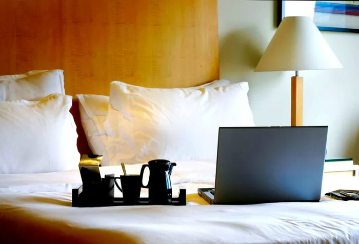 A laptop alongside a black coffee cup rests on a white bed with a wooden headboard.