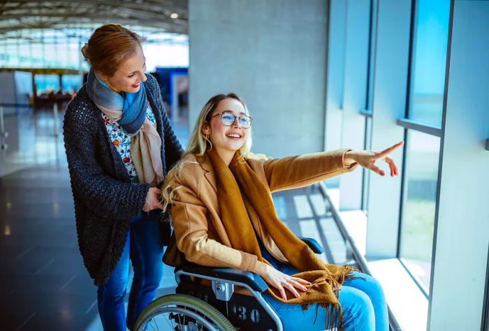 A woman assists a young woman in a wheelchair as they navigate through an airport terminal.