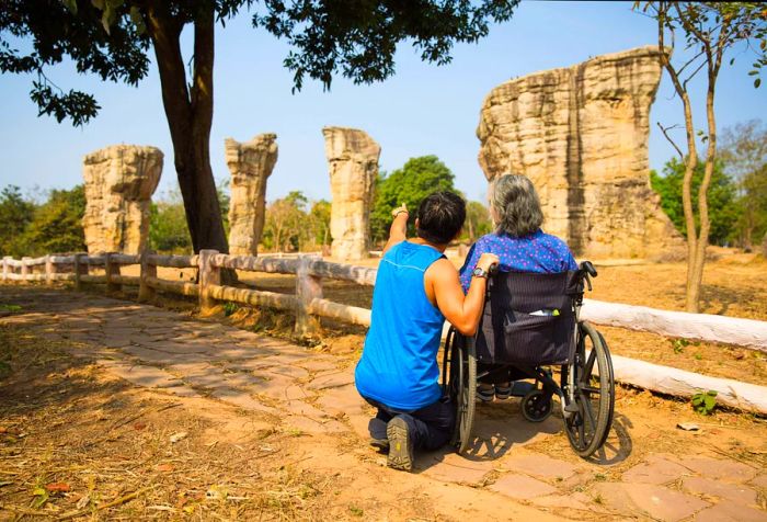 A man in a blue tank top and black athletic pants pushes his elderly mother in a wheelchair to admire the unique rock formations of Mo Hin Khao (often referred to as Thailand's Stonehenge) in Chaiyaphum Province, Thailand.