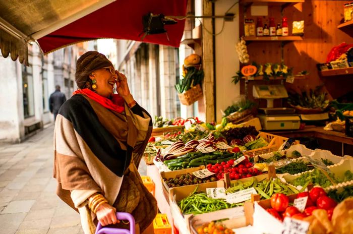 An elderly woman, bundled up in winter attire and wearing a turban, beams at a grocery market stall in Venice.