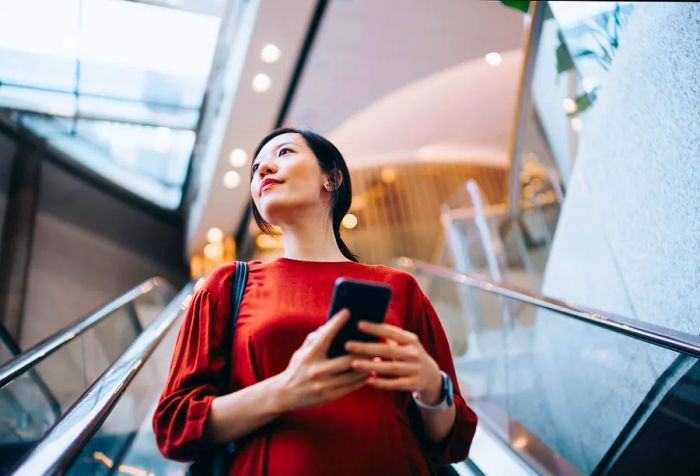 A young Asian woman confidently holds her smartphone with both hands while riding an escalator, looking thoughtfully into the distance.