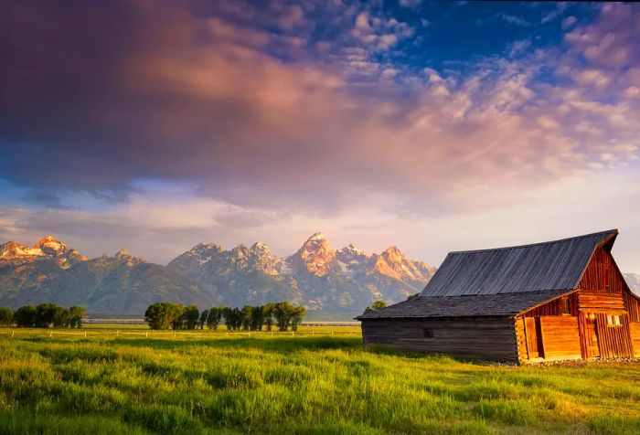 An old wooden barn set amidst lush ranchland, framed by a dramatic, snow-capped mountain range.