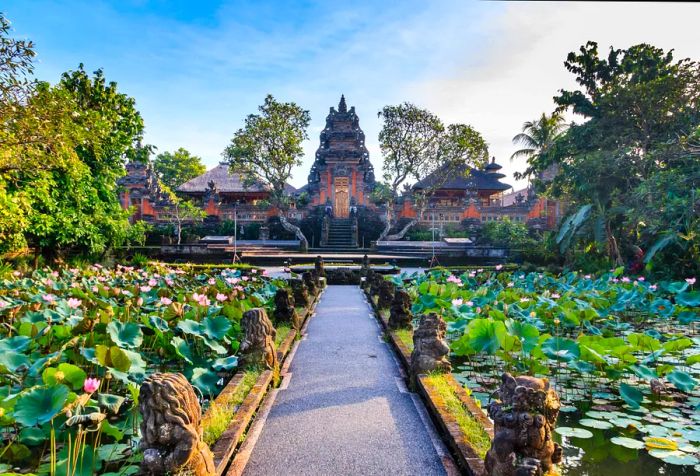 A walkway adorned with mythical creatures leads through a lotus garden to a grand entrance flanked by two cottages, featuring a tall gateway with golden doors.