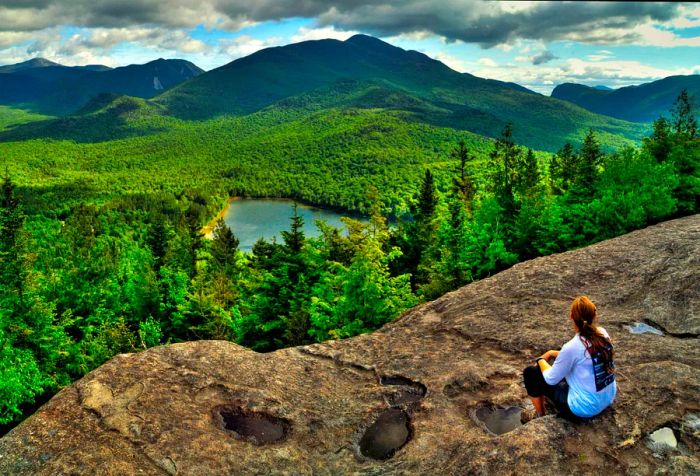 A person enjoys a view from a cliff, overlooking lush green hills and a picturesque lake nestled in the valley below.