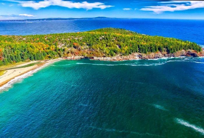 The deep blue ocean crashing against the sandy shore and rocky edges of a forested island.