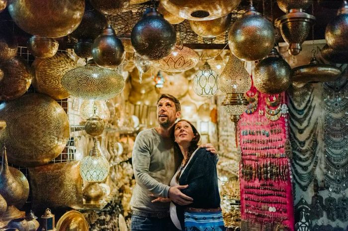 A couple stands smiling in a lamp shop in Marrakech, with the woman pregnant, both captivated by the enchanting lights.