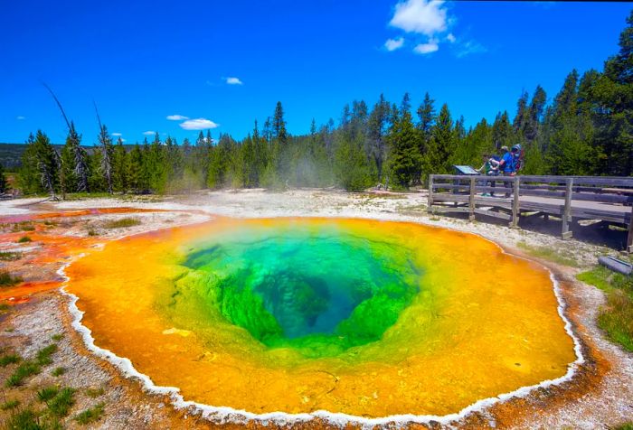 Two people stand on a viewing deck, gazing at a hot spring with vibrant colors caused by bacteria.