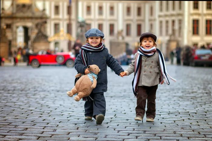 Two young brothers walk hand in hand down a cobblestone street in Old Town, Prague, with one boy clutching a stuffed bear.