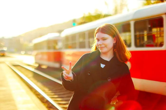 A teenage girl stands in front of an old-fashioned tram, waving goodbye with a smile.