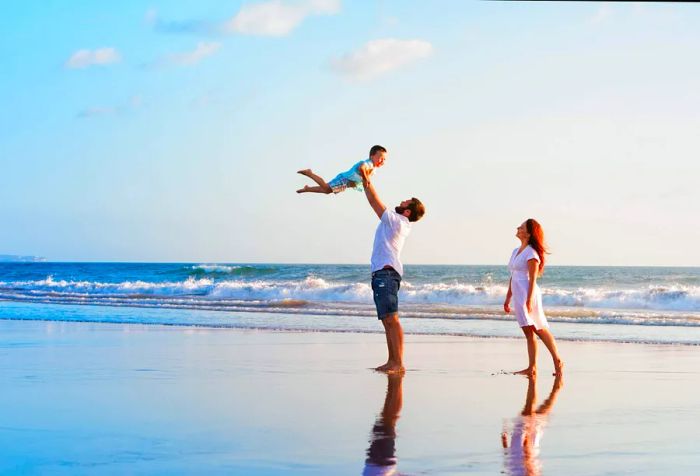 A man playfully lifting a young boy into the air while a woman observes from the beach behind them.