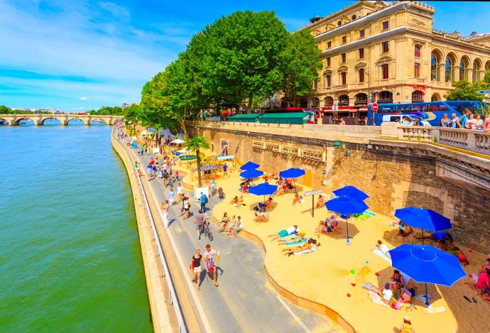 A riverside walkway features a sandy area where people unwind beneath parasols.