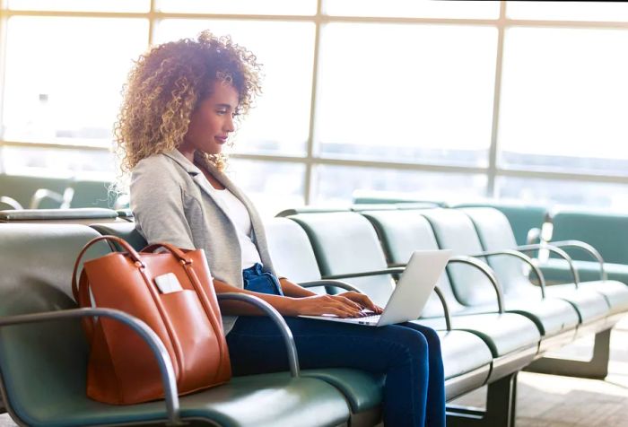 A woman sits at the airport, focused on her laptop, with her brown bag resting on a chair beside her.