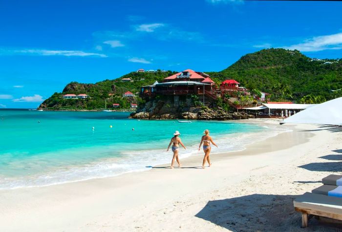 Two women in bikinis leisurely walking along a powdery white sand beach.