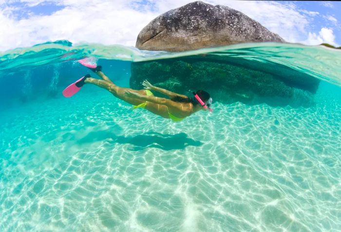 A woman in a vibrant green bikini navigating around a massive sunken boulder in crystal-clear waters.