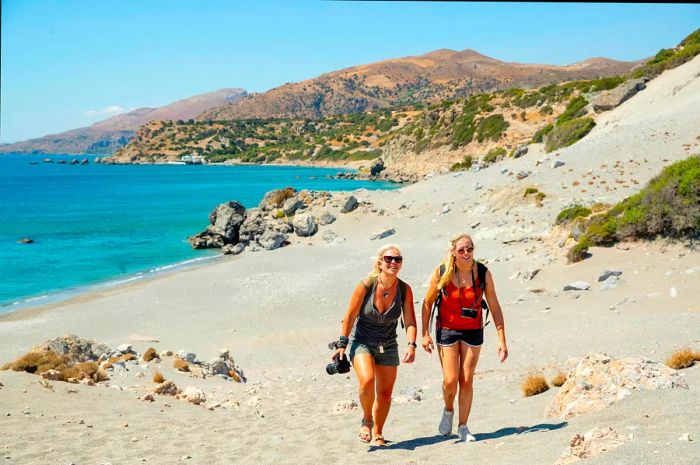 Two blonde women stroll along a beach in Crete, radiating happiness and carefree joy in the sun while capturing moments with their cameras.