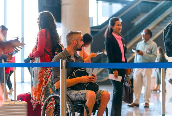 A disabled Caucasian male in a wheelchair is traveling, waiting in the check-in line at the airport.