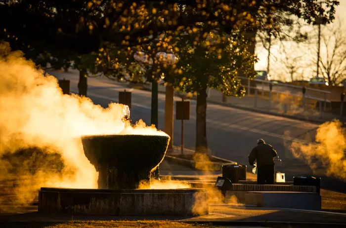 As the golden hour light filters through, steam gently rises from a fountain of hot spring water while a man fills his jugs in Hot Springs, Arkansas, creating a truly enchanting scene.