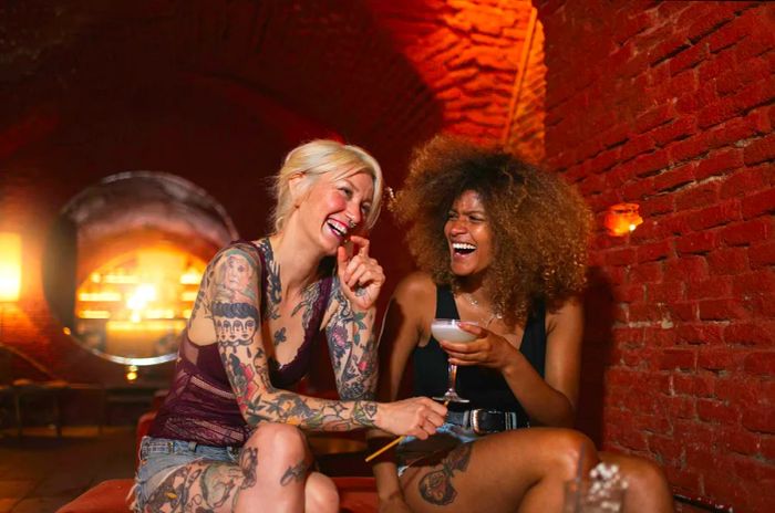 Two multiethnic female friends with tattoos laughing and enjoying alcoholic beverages while spending their weekend in a loft bar in Madrid, Spain